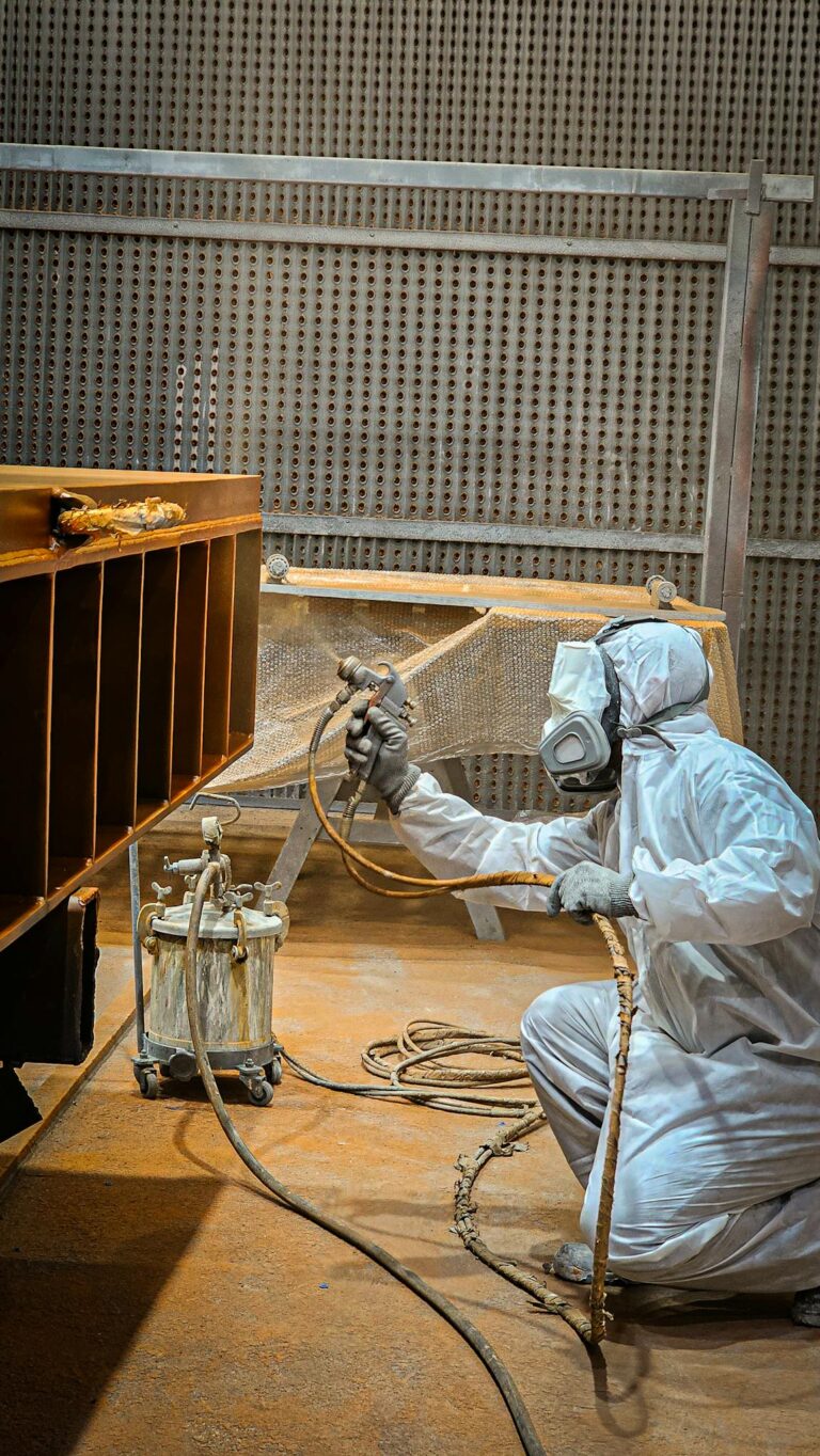 A worker in protective gear spray painting a metal structure indoors. Industrial setting.